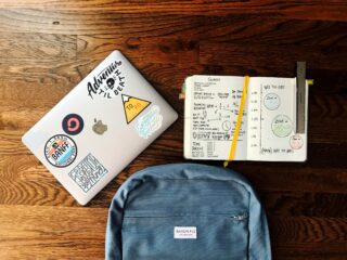 A backpack, laptop and an open journal with notes from class spread out on a wooden table.