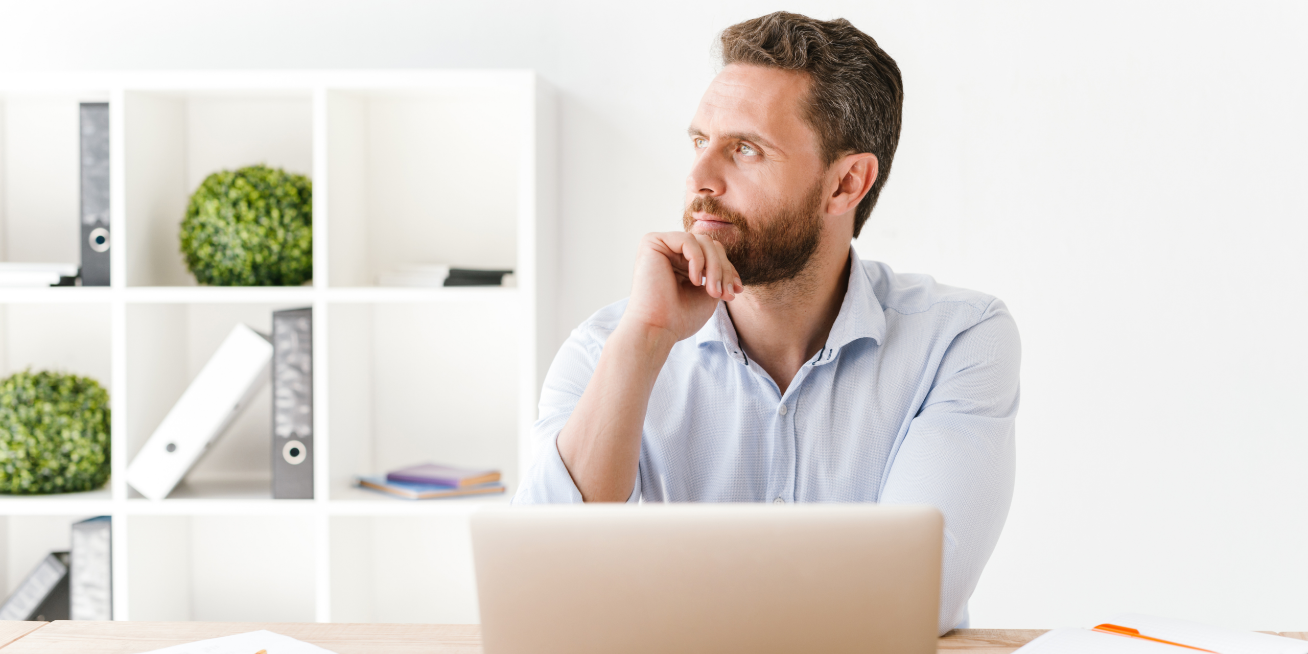 A professional man pausing to think while working at his desk with a laptop, notebooks, and a smartphone in a clean, minimalist workspace.